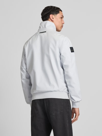 Männermodel trägt Bomberjacke mit Stehkragen Modell 'RACING' von Helly Hansen in hellem Grau, mit Logo-Print und Gerippte-Abschlüsse, Ganzkörperansicht Rückansicht. Rückenlänge bei Größe M: 68 cm, Schulterbreite bei Größe M: 43 cm, Brustweite bei Größe M: 58 cm, Ärmellänge bei Größe M: 68 cm. Gerader-Schnitt, Seitliche Eingrifftaschen, Reißverschluss.