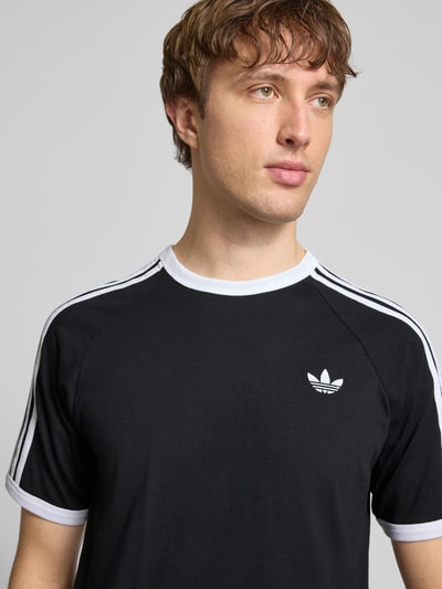 Männermodel trägt T-Shirt mit Label-Stitching von adidas Originals in Schwarz-Weiß mit Rundhalsausschnitt, Nahaufnahme Oberkörper.