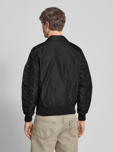 Männermodel trägt schwarze Regular Fit Bomberjacke mit Logo-Patch von Alpha Industries, elastische Abschlüsse, Ganzkörperansicht Rückansicht.