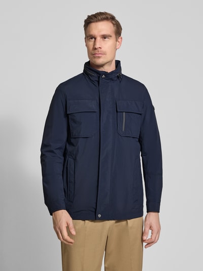 Męski model nosi Regular Fit Jacke mit Stehkragen od Bugatti, granatowa kurtka z kieszeniami na piersi, aplikacją z logo, krój regular fit, kaptur chowany, długość tyłu w rozmiarze 50: 72 cm, szerokość ramion: 46 cm, szerokość w rozmiarze 50: 52 cm, długość rękawów: 68 cm, widok całej sylwetki z przodu.