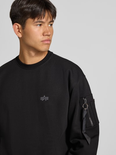 Męskąmodel nosi bluza 'Sweatshirt mit Label-Details' od Alpha Industries, mieszanka bawełny, luźny krój, okrągły dekolt, kieszeń na rękawie z aplikacją z metką, prążkowane wykończenia, kolor czarny, tekstura gładka, widoczne logo, długość tyłu 66 cm, długość rękawów 62 cm, zbliżenie na górną część ciała, widok z przodu.