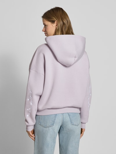 Frauenmodel trägt Relaxed Fit Hoodie mit Logo-Print von Review in Flieder, florales Stitching, Känguru-Tasche, Rückansicht Ganzkörperansicht.