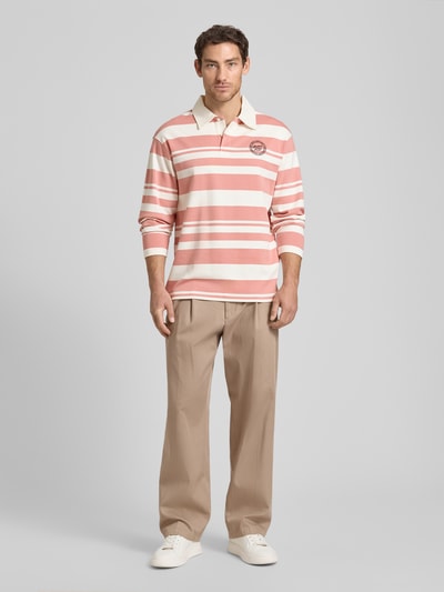 Męski model nosi koszulkę polo Relaxed Fit Poloshirt aus reiner Bio-Baumwolle Modell 'JACK' od SELECTED HOMME, efekt: wzór na całej powierzchni, krój relaxed fit, aplikacja z wyhaftowanym logo, kołnierzyk polo, zapięcie na 2 guziki, długi rękaw, wykończenie materiału: dzianina dresowa, materiał: bawełna ekologiczna, kolory różowy i kremowy, jasnobeżowe spodnie, białe buty, całkowity widok z przodu.