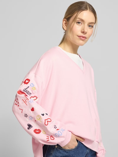 Vrouwenmodel draagt Oversized Fit Sweatshirt mit V-Ausschnitt van Smith and Soul in roze met kleurrijke motieven, katoen-modalmix, elastische boorden, vooraanzicht, bovenlichaam close-up.