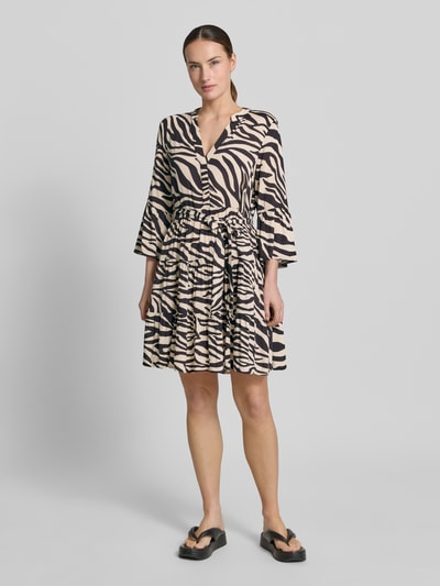 Vrouwenmodel draagt Tailliertes Tunikakleid aus reiner Viskose Modell 'MELBA' van Vero Moda met allover-print, tailliert fit, mini-lengte, V-hals, bindceintuur, trompetmouwen en volantzoom, natuurlijke kleuren, lichte textuur, vooraanzicht volledige lichaamsopname.