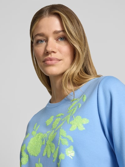 Frauenmodel trägt T-Shirt aus Viskose-Mix mit Paillettenbesatz von Milano Italy, hellblau mit gelbem Motiv-Print, Nahaufnahme Oberkörper.
