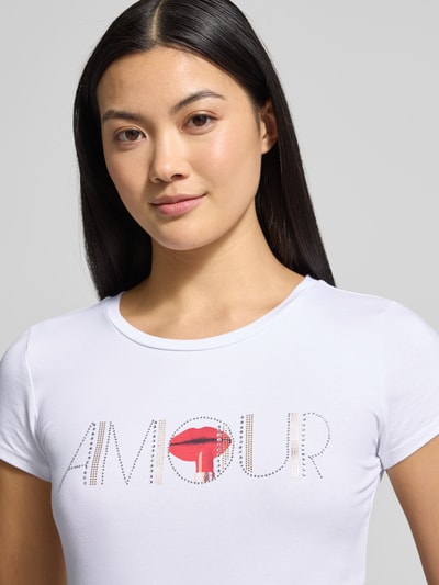 Kobiece model prezentuje T-shirt z Ziersteinbesatz od Guess, biały, slim fit, bawełna z elastanem, okrągły dekolt, z przodu napis, zbliżenie na górną część ciała.