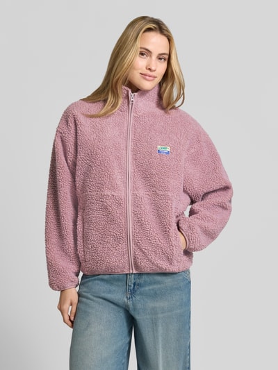 Frauenmodel trägt Regular Fit Jacke aus Teddyfell Modell 'Hoktown' von American Vintage in Rosa mit Stehkragen und Logo-Patch, Nahaufnahme Oberkörper. Schulterbreite 54 cm, Brustweite 46 cm, Rückenlänge 62 cm, Ärmellänge 55 cm.
