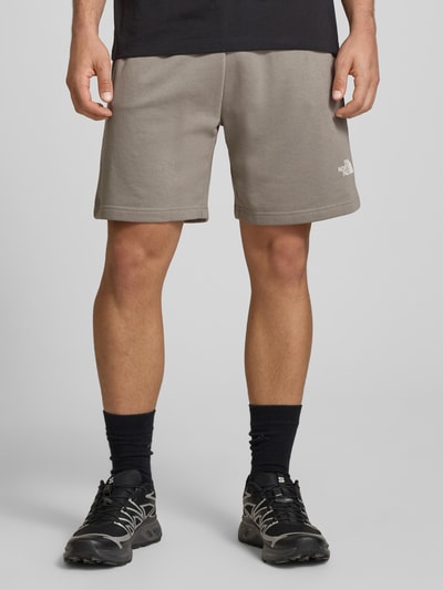 Mężczyzna nosi Shorts mit Logo od The North Face, bawełniany miks, krój regular fit, elastyczny pas, szerokość w rozmiarze M 41 cm, długość nogawki od wewnątrz w rozmiarze M 18 cm, jasnoszary, logo na nogawce, tekstura gładka, całkowity widok z przodu