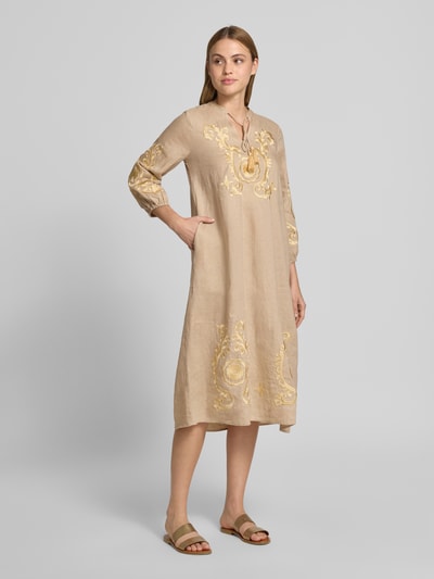 Frauenmodel trägt Tunikakleid aus Leinen mit Stitchings von Emily Van den Bergh, Gerader-Schnitt, Midikleid, Schnürung, 3/4-Arm, Ganzkörperansicht von vorne. Rückenlänge bei Größe 36: 117 cm, Brustweite: 37 cm, Ärmellänge: 51 cm, beige mit gelben Stickereien, glatte Textur.