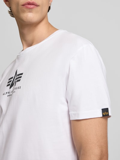 Männermodel trägt T-Shirt mit Logo und Rundhalsausschnitt van Alpha Industries in wit, katoen, regular fit, logo op borst, detailopname bovenlichaam.