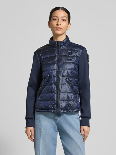 Frauenmodel trägt Steppjacke mit Label-Patches Modell 'HASKEL' von Blauer Usa in Dunkelblau, glänzende Textur, Ganzkörperansicht von vorne.
