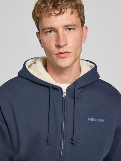 Männermodel trägt Regular Fit Sweatjacke aus Baumwoll-Mix mit Teddyfutter von REVIEW, dunkelblau, Logo-Detail, Nahaufnahme Oberkörper.