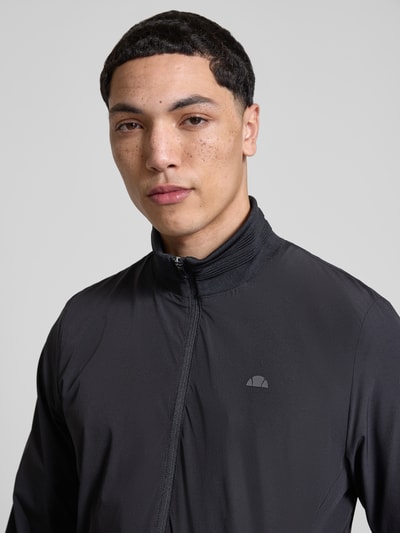 Männermodel trägt Sweatjacke mit Logo-Print Modell 'CANDORA' von Ellesse in Schwarz, glatte Textur, Nahaufnahme Oberkörper. Gerader Schnitt, Stehkragen und Reißverschluss aus Material-Mix mit Stretch-Anteil deuten auf hohen Tragekomfort hin.