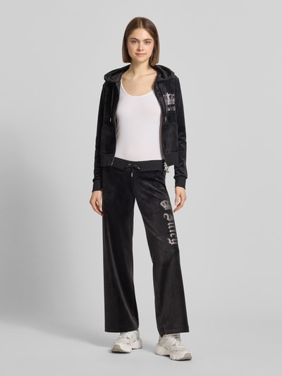 Kobiece modelka nosi czarną Sweatjacke mit Ziersteinbesatz Modell 'GOTHIC CROWN ROBERTSON' od Juicy Couture z kapturem, krój slim fit, materiał z domieszką stretchu, dwukierunkowy zamek, widoczne zdobienia z kamieni, tkanina miękka, gładka faktura, widok całej sylwetki z przodu.