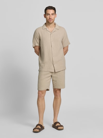 Męski model nosi szorty z wpuszczanymi kieszeniami i elastycznym pasem od Lindbergh, beżowa bawełna, krój loose fit, szerokość w rozmiarze M 43 cm, długość wewnętrzna nogawki 22 cm, widok całej sylwetki z przodu.