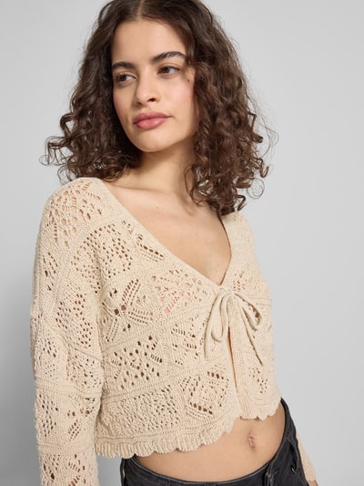 Modelka nosi Cropped Strickjacke aus Baumwoll-Mix Modell 'BEACH LIFE' od Only, kolor jednolity, krój relaxed fit, wzór ażurowy, dekolt V, długi rękaw z falistym brzegiem, przędza bawełniana, widok na górną część ciała z przodu.