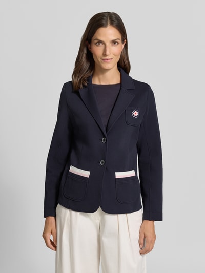 Vrouwenmodel draagt Regular Fit Blazer met Reverskraag van Marc Cain, katoenmix met stretch, borstzak met bloemenapplicatie, opgestikte zakken, knoopsluiting, kleur donkerblauw, zakdetails in wit en roze, lengte 59 cm, borstwijdte 47 cm, mouwlengte 63 cm, frontale opname van het hele lichaam.