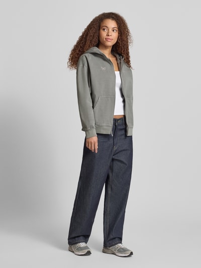 Modelka nosi Oversized Sweatjacke mit Känguru-Tasche Modell 'NIVELLES' od Pegador, szara bawełniana bluza z logo, zamek, kieszeń; widok całej sylwetki z przodu.