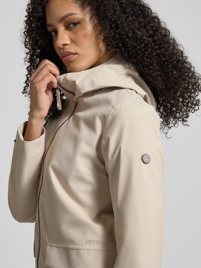Frauenmodel trägt Regular Fit Parka mit Logo-Applikation Modell 'Folka' von Didriksons in Beige mit Stehkragen, Kapuze und Zweiwege-Reißverschluss, aufgesetzte Taschen, Rückenlänge 83 cm, Schulterbreite 40 cm, Brustweite 46 cm, Ärmellänge 64 cm, Nahaufnahme Oberkörper.