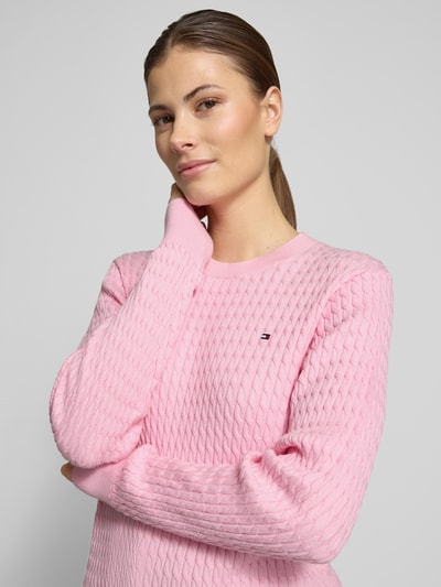 Frauenmodel trägt Slim Fit Pullover aus reiner Baumwolle von Tommy Hilfiger in Rosa mit Zopfmuster, Rundhals, Strickoptik, Nahaufnahme Oberkörper.