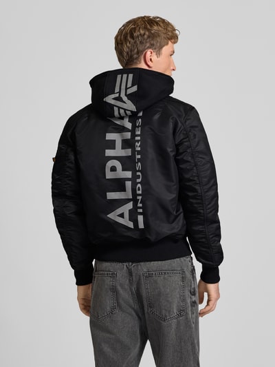 Männermodel trägt Regular Fit Bomberjacke mit Kapuze von Alpha Industries in Schwarz mit großem Label-Patch, Ganzkörperansicht Rückansicht. Herren Bomberjacke aus Webstoff, langärmliges Design, durchgängiger Zipper, gerippte Abschlüsse, Eingrifftaschen, Armtasche mit Label-Detail, gerippter Stehkragen.