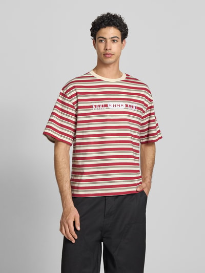 Męski t-shirt z bawełny w czerwone i beżowe paski, Relaxed Fit, model 'Heritage', wyhaftowane logo KARL KANI, okrągły dekolt, długość tyłu w rozmiarze M: 64 cm. Mężczyzna nosi Relaxed Fit T-Shirt mit Logo-Stitching Modell 'Heritage' od KARL KANI, Ganzkörperansicht von vorne.