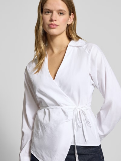 Modelka kobieca ma na sobie Regular Fit Hemdbluse mit Umlegekragen od Gina Tricot, biała bawełna, krój regular fit, dopasowana talia, długi rękaw, dekolt V, asymetryczny fason, widok na całą sylwetkę z przodu.