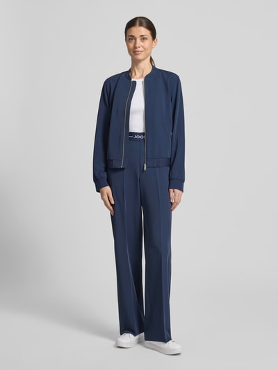 Kobiece model prezentuje spodnie Hose mit elastischem Bund und Label-Detail von JOOP!, mieszanka materiałów z dodatkiem streczu, krój regular fit, elastyczny pas, detal z logo, szerokość w rozmiarze 36: 38 cm, długość wewnętrzna nogawki w rozmiarze 36: 81 cm, granatowy kolor, prosta nogawka, gładka tekstura, widok całej sylwetki z przodu.