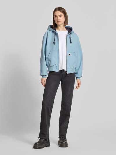 Frauenmodel trägt hellblaue Sweatjacke mit Kapuze und Känguru-Tasche von Carhartt Work In Progress aus Reine Baumwolle, Relaxed-Fit, mit Reißverschluss, Label-Detail, kombiniert zu schwarzer Jeans und schwarzen Schuhen, Ganzkörperansicht von vorne. Rückenlänge bei Größe S: 59 cm, Schulterbreite: 52 cm, Brustweite: 58 cm, Ärmellänge: 60 cm.