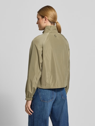 Modelka kobieca ma na sobie kurtkę 'Jacke mit elastischen Abschlüssen und Stehkragen' od Fuchs Schmitt, kolor oliwkowy, prosty krój, stójka, zamek błyskawiczny, raglanowe rękawy, boczne kieszenie zapinane na zamek błyskawiczny, elastyczne wykończenia, długość tyłu w rozmiarze 36: 56 cm, szerokość ramion w rozmiarze 36: 38 cm, szerokość w biuście w rozmiarze 36: 48 cm, długość rękawów w rozmiarze 36: 43 cm, tekstura gładka, widok całej sylwetki z tyłu.