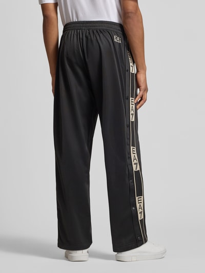 Männermodel trägt schwarze Trainingshose mit seitlichen Knopfleisten von EA7 Emporio Armani, Relaxed-Fit, Label-Details, Ganzkörperansicht Rückansicht.