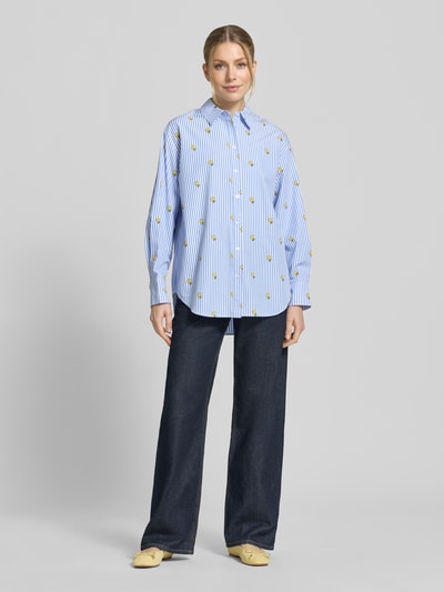 Modelka kobieca nosi Oversized Hemdbluse aus reiner Baumwolle Modell 'JEPPERMINT SNOOPY' od Essentiel Antwerp, niebiesko-białe paski, żółty nadruk, długie rękawy, popelina, widok całej sylwetki z przodu.