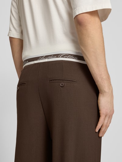 Männermodel trägt Stoffhose mit Logo-Bund und Gesäßtaschen von ANOTHER COTTON LAB, braun, Material-Mix mit Stretch-Anteil, Detailaufnahme Rückansicht.