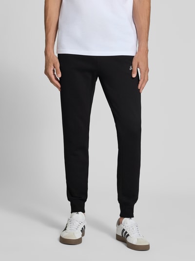 Męski model nosi spodnie dresowe Sweatpants mit Label-Print Modell 'GORDON' od JACK & JONES, czarne, wpuszczane kieszenie, luźny krój, widok z przodu, kontekst: elastyczny pas z tunelem, długość wewnętrzna nogawki w rozmiarze M: 77 cm. Ganzkörperansicht von vorne.