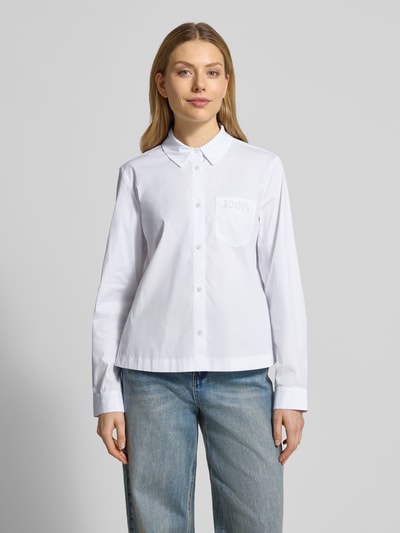 Modelka kobieca ma na sobie Regular Fit Hemdbluse mit Brusttasche Modell 'Bella' od JOOP!, biały, długi rękaw, kołnierz koszulowy, bawełna elastyczna, kieszeń na piersi, wyszywane logo; widok całej sylwetki z przodu.