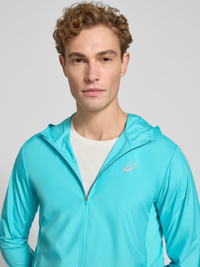 Männermodel trägt Windbreaker mit Logo-Print und Kapuze von ASICS in Türkis, glatte Textur, sichtbarer Reißverschluss, Nahaufnahme Oberkörper.