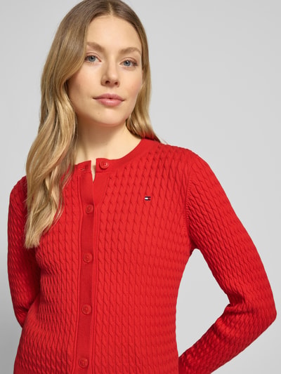 Frauenmodel trägt Slim Fit Strickjacke aus reiner Baumwolle von Tommy Hilfiger in Rot, Optik: Unifarben, Zopfmuster, Logo-Stitching, Rundhals, Langarm, Ärmelabschluss und Rumpfabschluss gerippt, Materialverarbeitung: Strick, Nahaufnahme Oberkörper.
