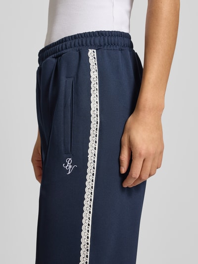 Kobiece model prezentuje Sportliche Trackhose mit geradem Bein od REVIEW, granatowy kolor, koronkowe elementy przy bocznym szwie, zaszewka z przodu, wyhaftowane logo na bocznej kieszeni, długość wewnętrzna nogawki w rozmiarze S: 39 cm, detaliczne ujęcie bocznego szwu.