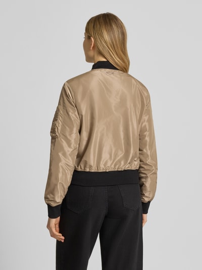 Frauenmodel trägt Bomberjacke mit Label-Patch und Reißverschlusstasche am Ärmel von Original Bombers in Beige, schwarze Abschlüsse, Lockerer-Schnitt, Stehkragen, Rückenlänge 53 cm, Schulterbreite 37 cm, Brustweite 48 cm, Ärmellänge 63 cm, Ganzkörperansicht Rückansicht.