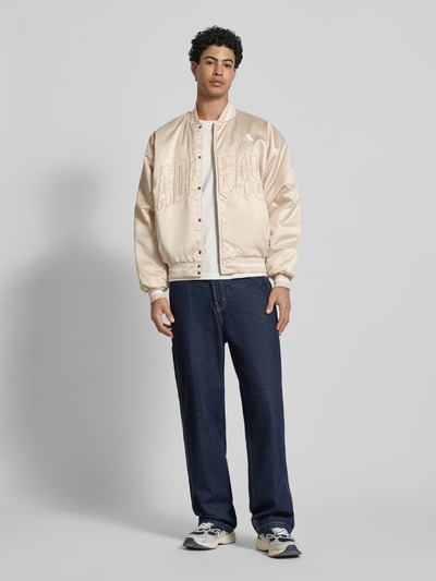 Männermodel trägt Regular Fit Bomberjacke mit Stehkragen von adidas Sportswear, cremefarben, Logo-Stitching, Ganzkörperansicht von vorne. Rückenlänge bei Größe M 69 cm, Schulterbreite 64 cm, Brustweite 67 cm, Ärmellänge 61 cm, elastische Abschlüsse, Druckknopfverschluss.