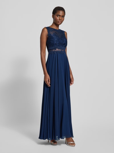 Frauenmodel trägt Abendkleid mit floralen Stickereien von Mascara in Dunkelblau, taillierte Passform, mehrlagiger Rockteil, rückenlanger Reißverschluss, transparente Einsätze, wattierte Cups, ärmellos, Ganzkörperansicht von vorne.