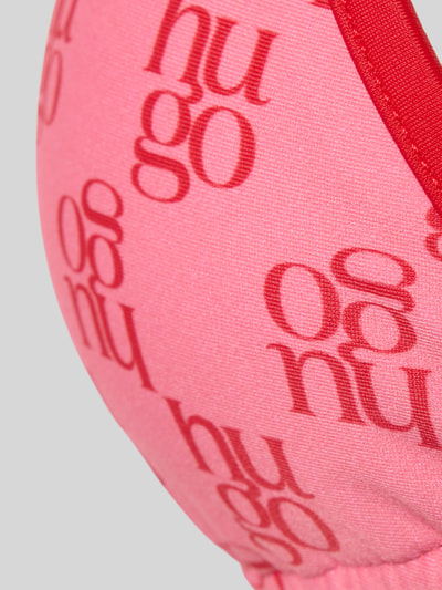 Frauenmodel trägt Bikini-Oberteil mit Logo-Muster von HUGO in Rosa mit roten Schriftzügen, strukturierter Stoff, Nahaufnahme Oberkörper.