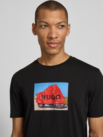 Männermodel trägt Relaxed Fit T-Shirt aus reiner Baumwolle Modell 'DOSTINO' von HUGO in Schwarz mit Label Print auf der Brust, Nahaufnahme Oberkörper.