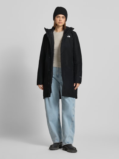 Vrouwenmodel draagt lange jas Model 'SUZANNE' van The North Face in zwart met recht pasvorm, capuchon, ritssluiting, labeldetail, zij-steekzakken, ruglengte maat S 92 cm, schouderbreedte 40 cm, borstwijdte 48 cm, mouwlengte 66 cm, geheel lichaamsaanzicht van voren.