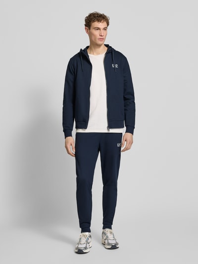 Männermodel trägt Slim Fit Sweatjacke mit Kapuze von EA7 Emporio Armani in Dunkelblau, elastischer Baumwoll-Mix, Ganzkörperansicht von vorne.