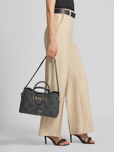 Kobiece model nosi Handtasche mit Logo-Muster Modell 'JANIE' od Guess, czarne logo, krótkie i regulowane rączki, Szerokość w rozmiarze 33 cm, długość 21 cm, głębokość 11 cm, długość paska 85 cm, detal z logo, widok całej sylwetki z przodu.