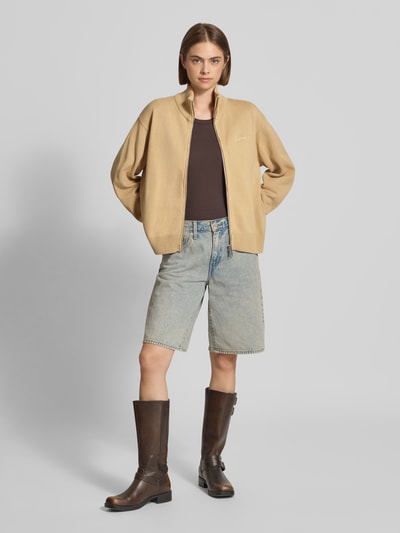 Frauenmodel trägt Cardigan mit Label-Stitching Modell 'Kori' von Carhartt Work In Progress in Beige, Stehkragen, Zweiwege-Reißverschluss, Baumwolle, Relaxed Fit, kombiniert mit Jeansshorts, Ganzkörperansicht von vorne.