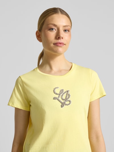 Kobiece modelka nosi t-shirt z bawełny w kolorze jasnej żółci, z prostym krojem i okrągłym dekoltem, detal z logo z obszyciem kamieni stras, długość tyłu w rozmiarze S: 60 cm, T-Shirt mit Rundhalsausschnitt und Strasssteinbesatz od LIU JO SPORT, zbliżenie na górną część ciała, przód
