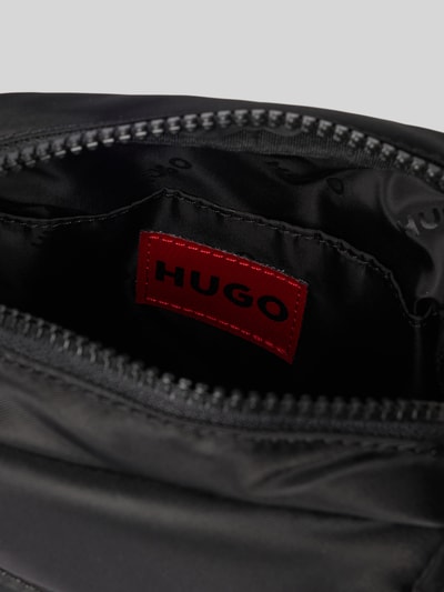 Produktfoto, czarny plecak z aplikacją z metką model 'AYDEN_MONOSTRAP' od HUGO, jednolity kolor, zamek błyskawiczny, główna przegroda, przegródka, przegródka zapinana na zamek błyskawiczny, możliwość noszenia na jedno ramię